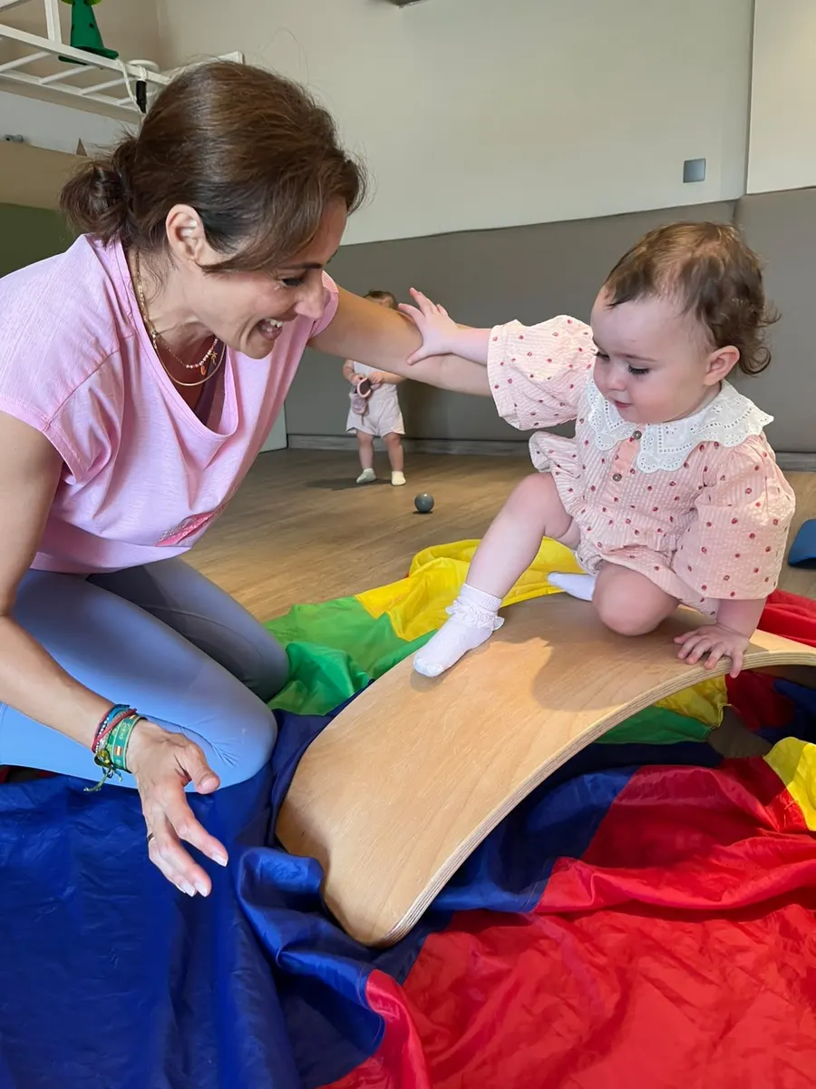 A teacher supporting a toddler balancing on a curved wooden board during a music and movement class