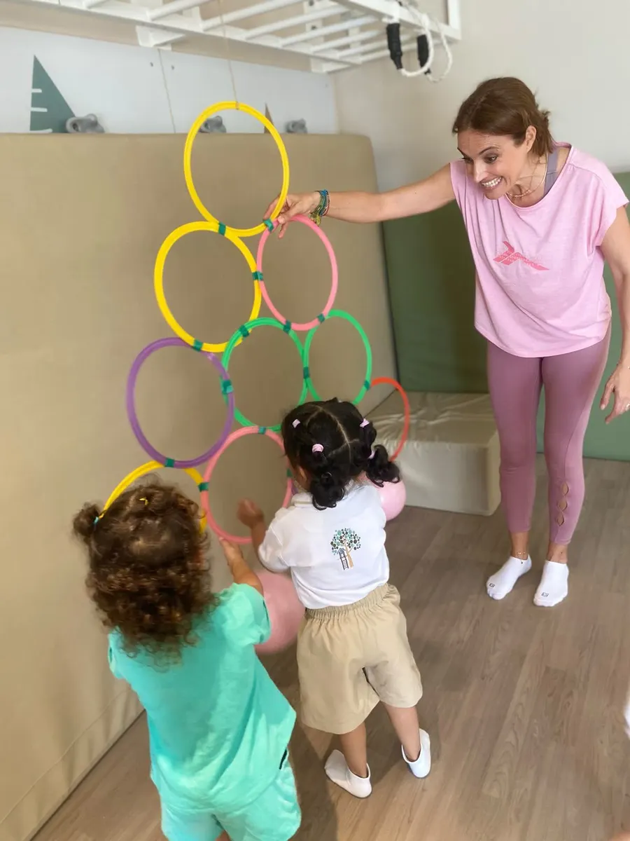 A teacher guiding two children reaching for coloured hoops on a padded wall in a music and movement class