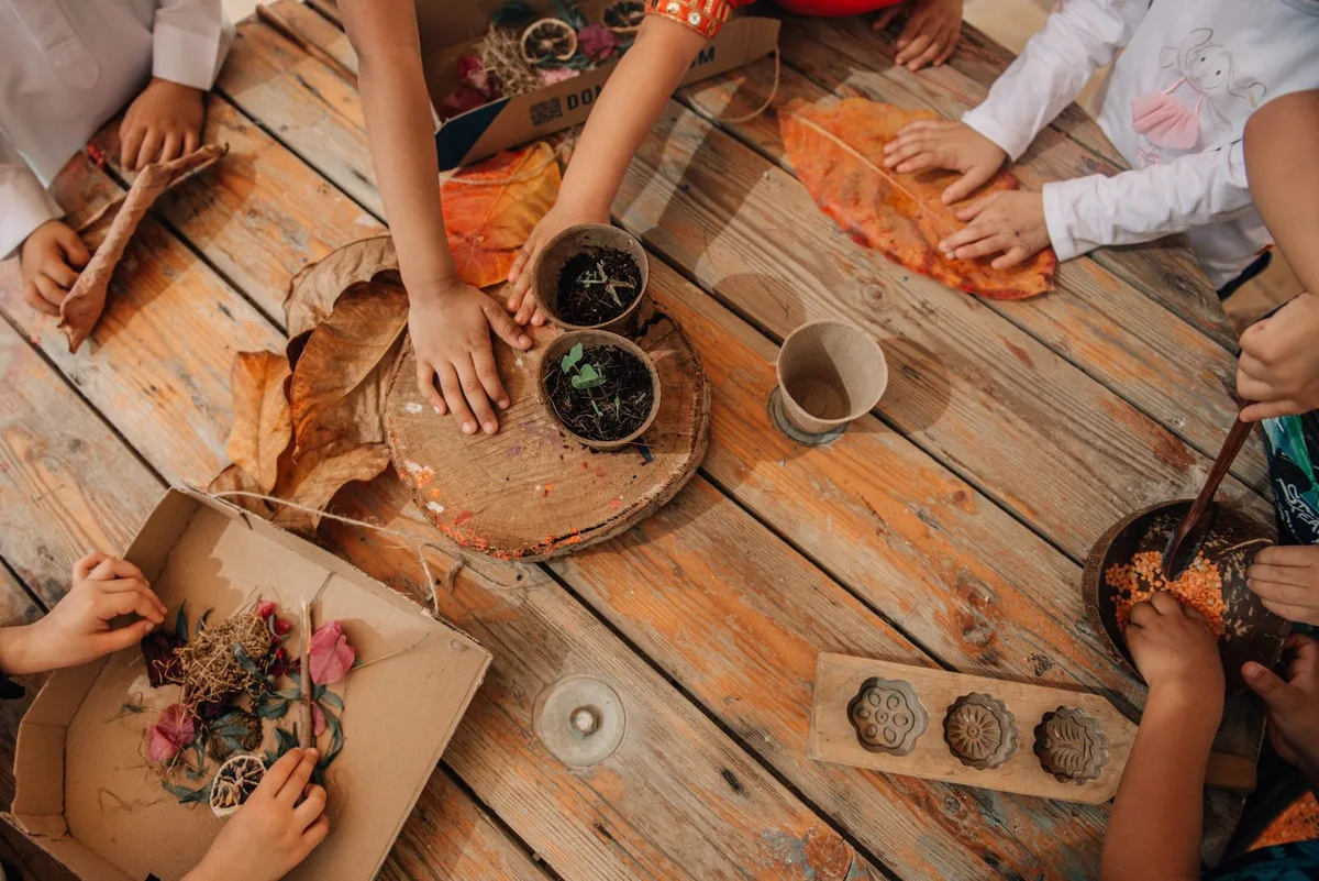 Children's hands arranging dried leaves, petals and seedling pots on a wooden outdoor table