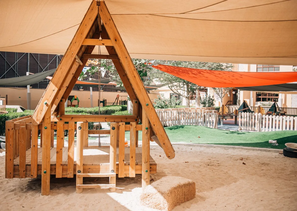 Wooden playhouse with swing over a shaded sand play area in the nursery garden