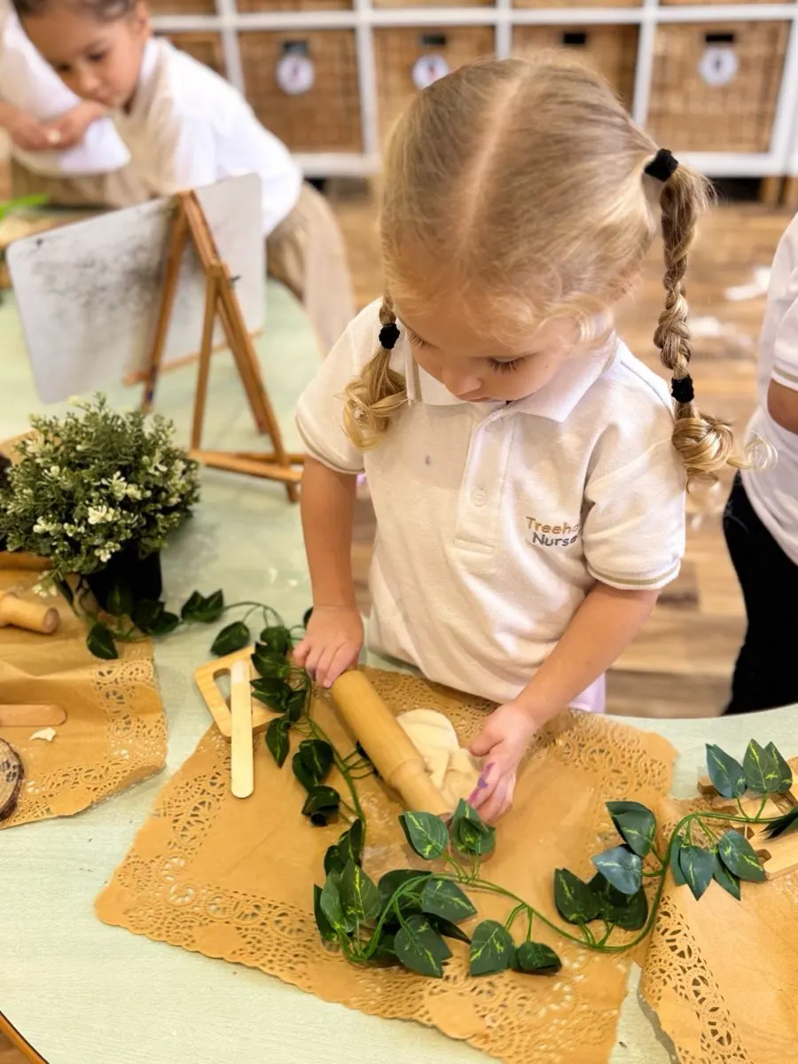 A child rolling dough beside green leaves and a small plant as part of a classroom nature project