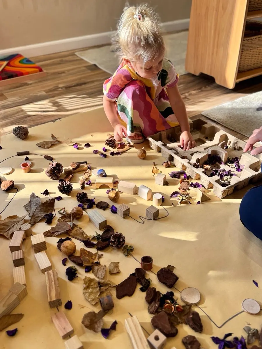 A child arranging pine cones, wood slices and dried flowers across a large floor tray