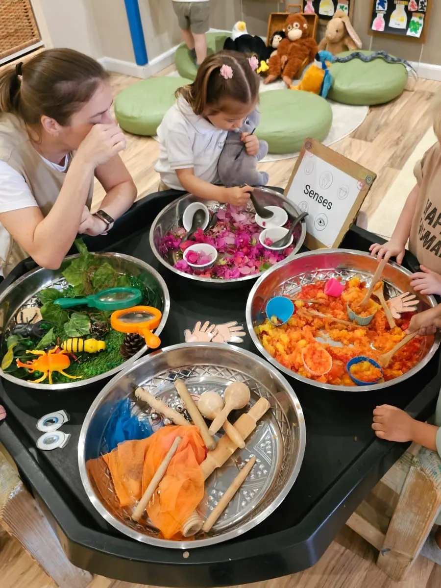 A teacher and children exploring trays of coloured sensory materials set out around a five senses sign